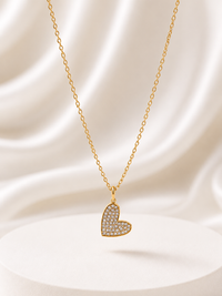 A gold heart-shaped pendant necklace paved with small clear stones, hanging from a delicate gold chain against a draped white satin background.