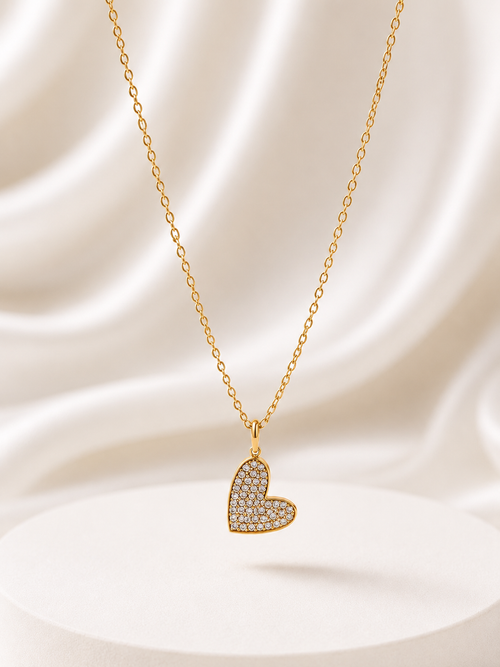 A gold heart-shaped pendant necklace paved with small clear stones, hanging from a delicate gold chain against a draped white satin background.