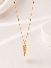 A delicate gold chain necklace featuring a textured wing pendant and small dark metallic bead accents, displayed on a soft, light beige background.