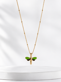 A gold chain necklace with a small dragonfly pendant featuring bright green wings and a gold body, displayed against a soft white background.