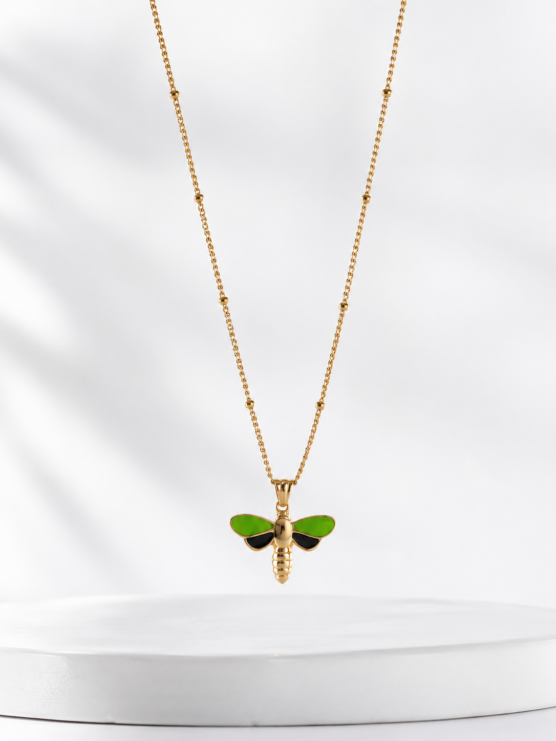 A gold chain necklace with a small dragonfly pendant featuring bright green wings and a gold body, displayed against a soft white background.
