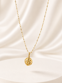 A delicate gold chain necklace with a textured leaf pendant, displayed against a draped cream satin fabric background.