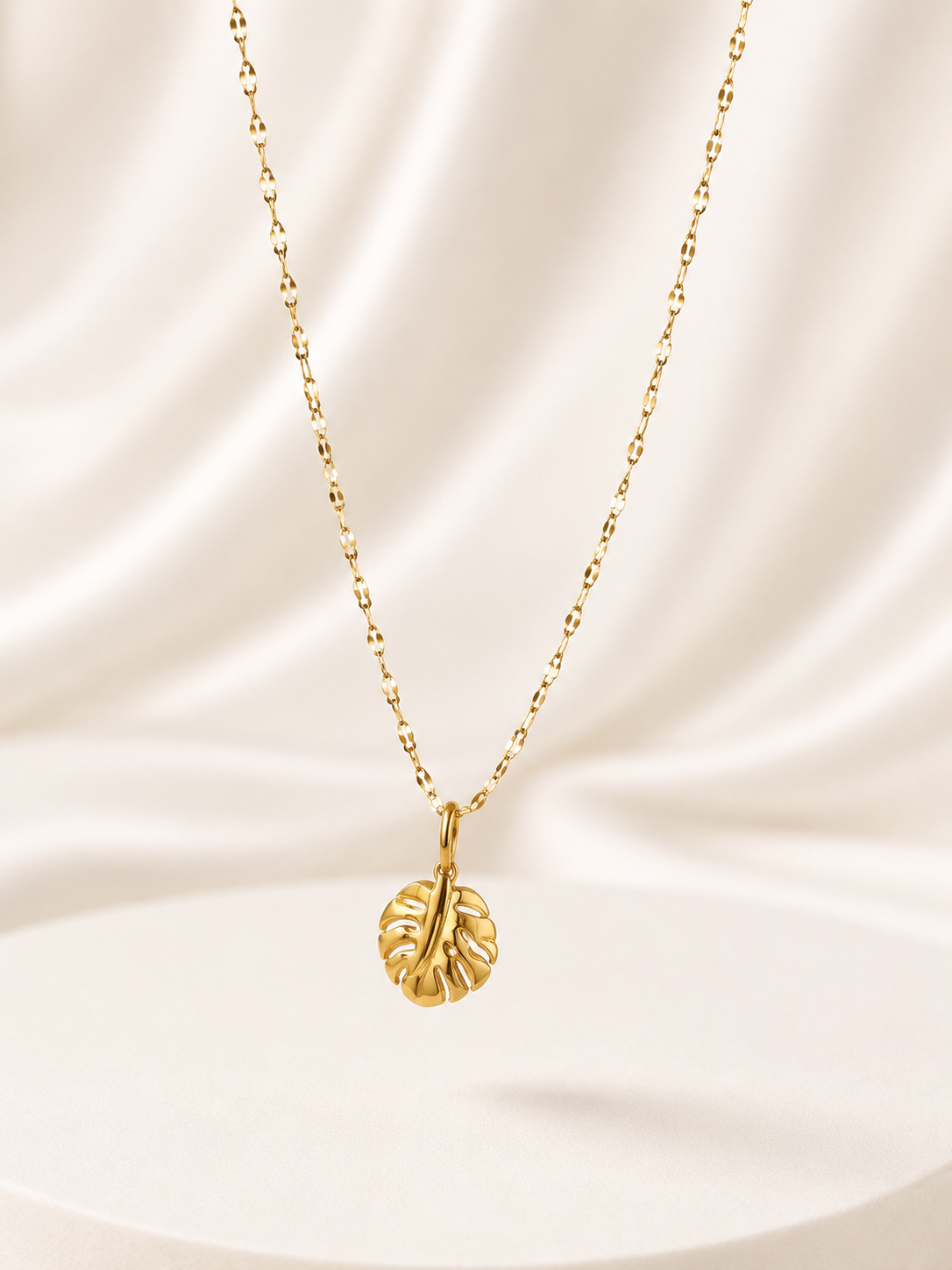 A delicate gold chain necklace with a textured leaf pendant, displayed against a draped cream satin fabric background.