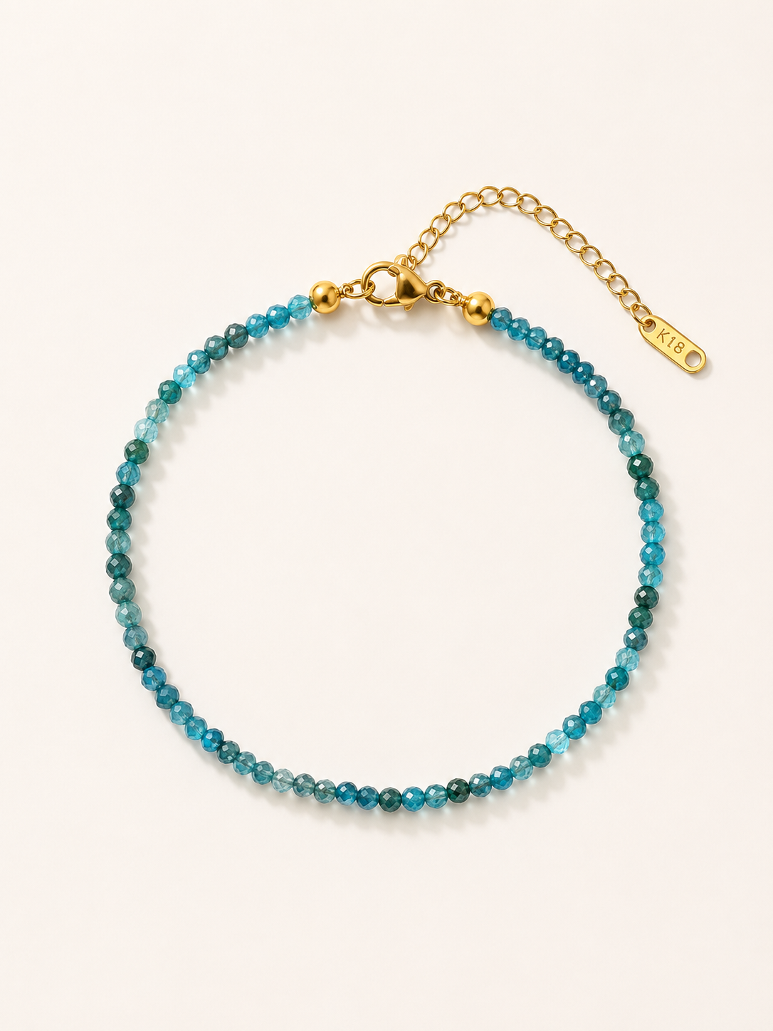 A beaded bracelet with a gradient of blue and green beads, accented by gold-tone hardware, including a lobster clasp, extension chain, and a small tag engraved with 'SILK'.