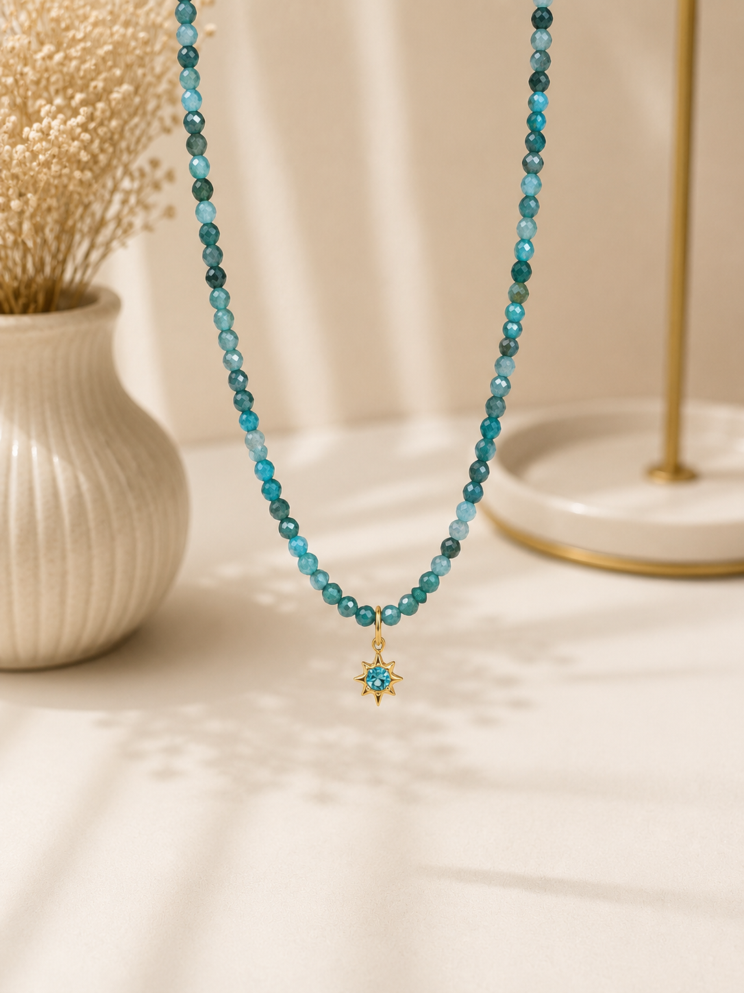 A beaded necklace with translucent teal-blue beads and a small gold-toned starburst pendant, displayed on a cream surface with a vase and dried flowers in the background.