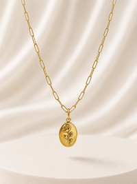 A gold paperclip chain necklace with an oval pendant engraved with a rose, displayed against a draped cream satin background.
