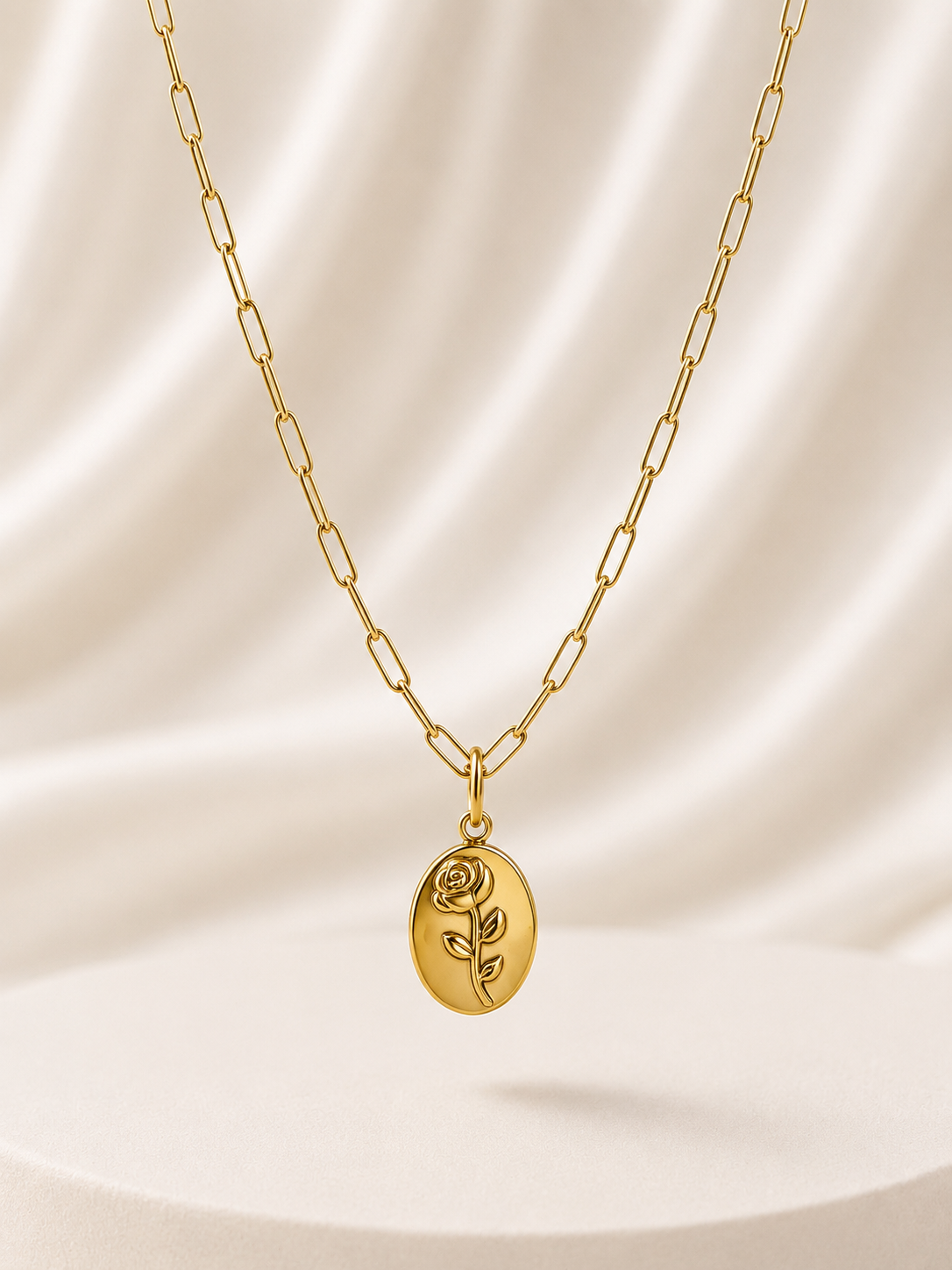 A gold paperclip chain necklace with an oval pendant engraved with a rose, displayed against a draped cream satin background.