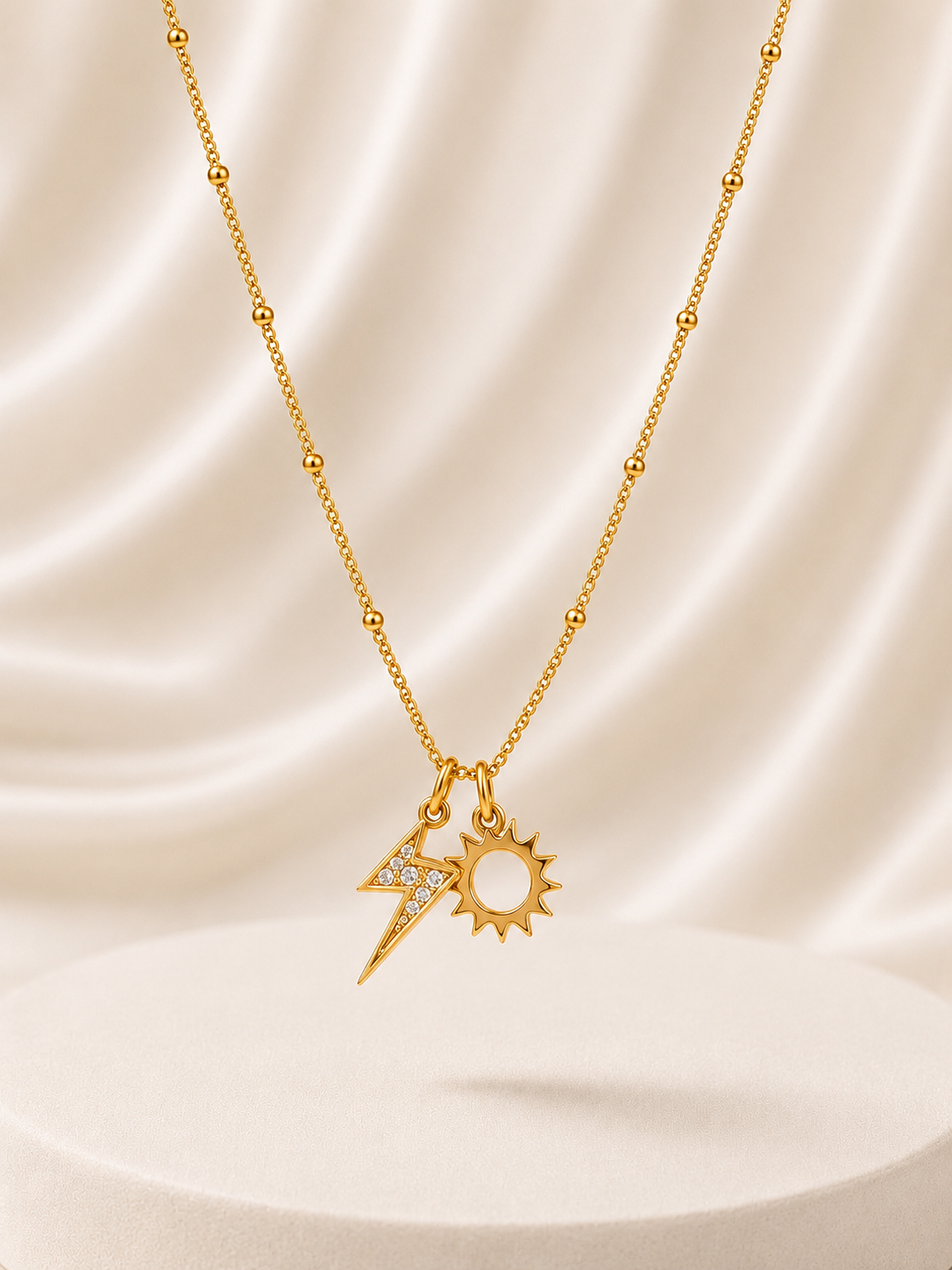 A gold necklace with a beaded chain and a double pendant featuring a lightning bolt and a sun, displayed on a cream satin drape background.