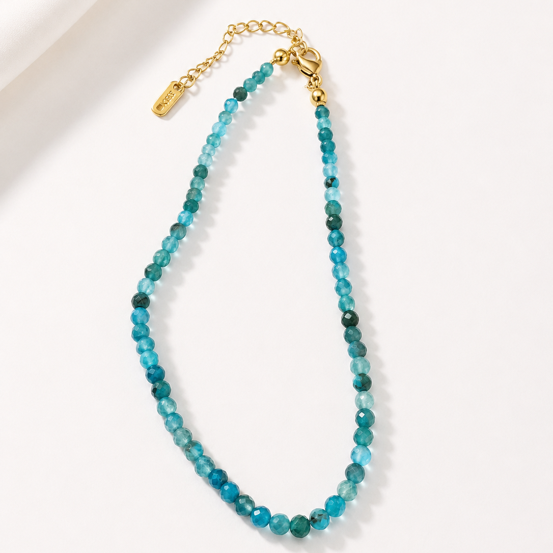 A beaded necklace made of faceted turquoise-blue beads with small dark green accents, finished with a gold-tone clasp and extender chain, photographed on a clean white background.
