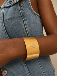 A close-up of a person wearing a wide, chunky gold cuff bracelet with a small diamond-encrusted cross on the front, paired with a blue denim top.