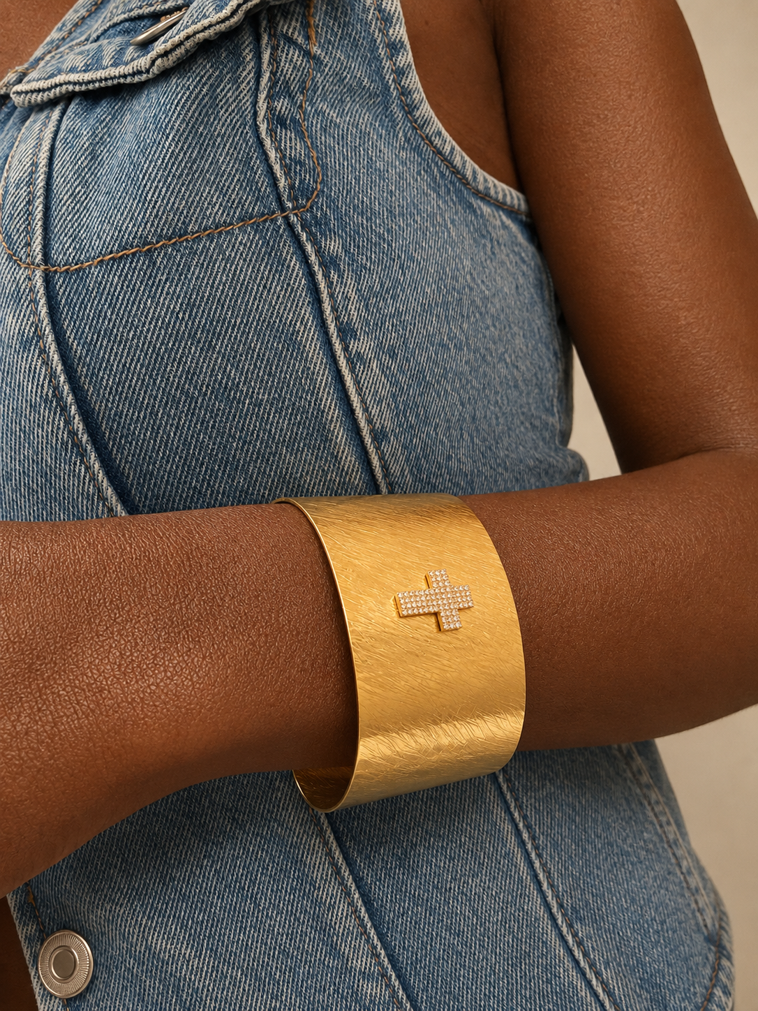 A close-up of a person wearing a wide, chunky gold cuff bracelet with a small diamond-encrusted cross on the front, paired with a blue denim top.