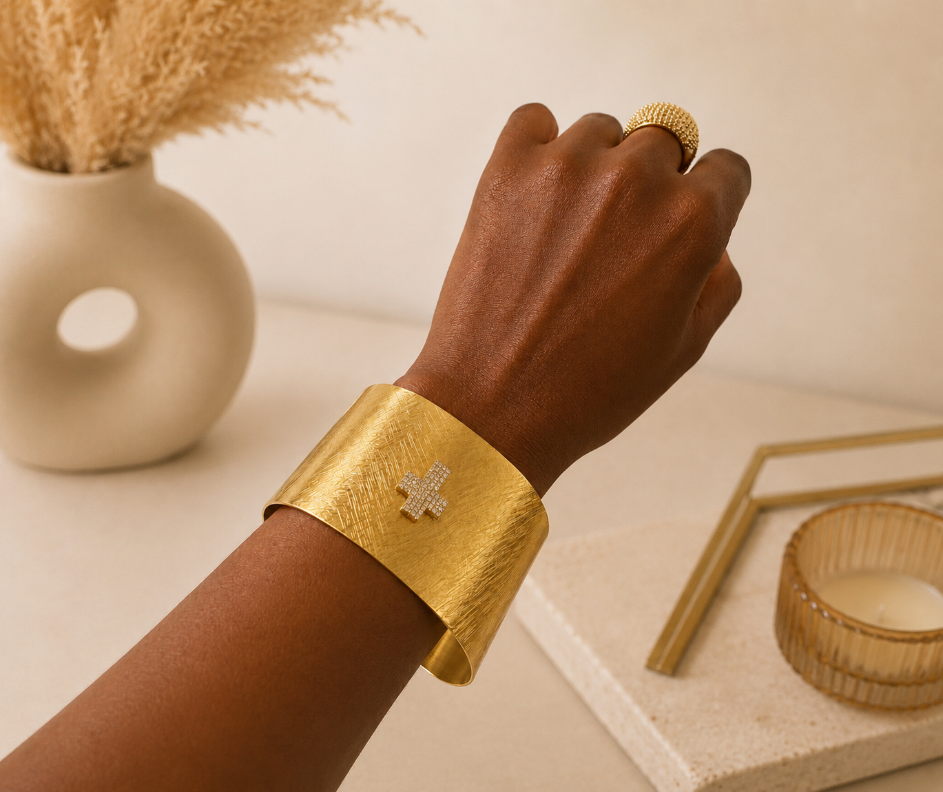 A close-up of a person's hand and wrist wearing a wide textured gold cuff bracelet with a small diamond-encrusted cross at the front, alongside a matching gold ring, set against a warm beige background with decorative dried grass and a candle.