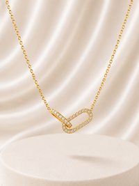 A gold chain necklace with an infinity-shaped pendant paved with small clear stones, displayed on a cream satin drape over a round pedestal.