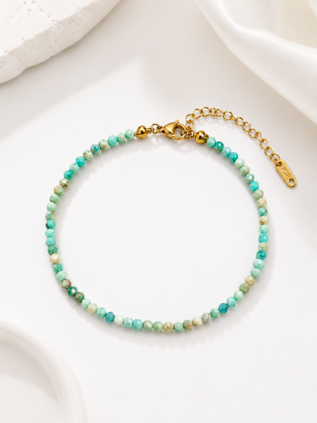 A delicate beaded bracelet with small turquoise and green beads, accented by gold-tone clasp and extender chain, displayed on a clean white surface.