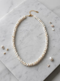 A double-strand pearl necklace with a gold clasp, arranged in an oval shape on a white marble background with small pearl and crystal accessories scattered around.